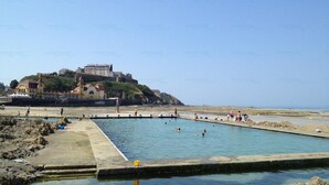 A coastal swimming area with a large pool, a beach, and a historic building on a hill in the background.