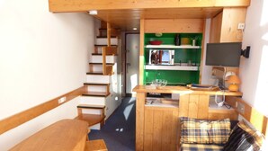 A compact kitchen with a microwave, stove, and sink. There is a wooden dining table with chairs and a small sofa with plaid cushions. A staircase leads to an upper level.