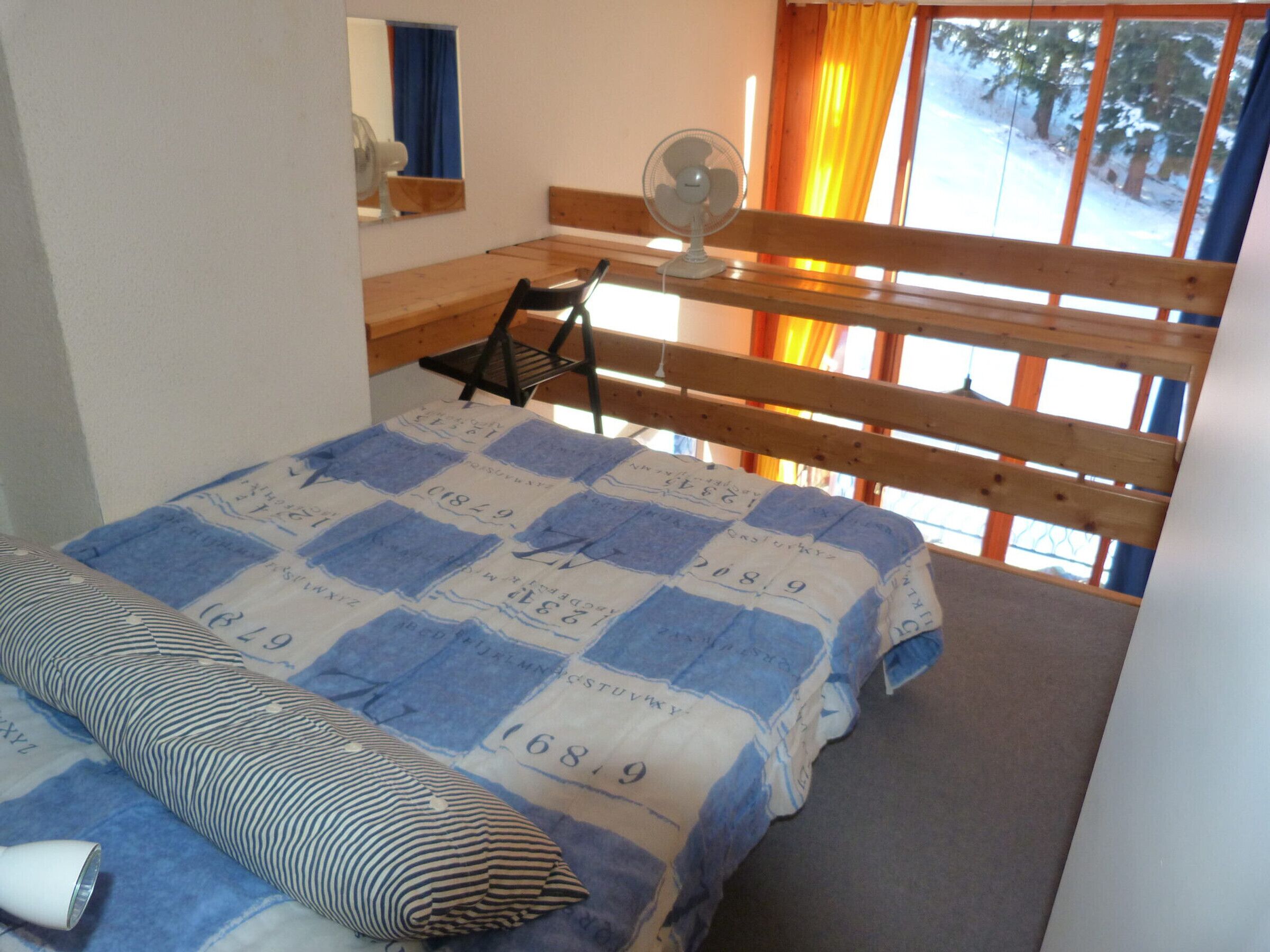 Ein Schlafzimmer mit einem Bett, einem Sessel, einem Ventilator und einem Fenster mit Vorhängen.