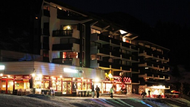 Ett hotell med flera våningar och balkonger, en restaurang på bottenvåningen och en skidbacke som ligger framför hotellet.