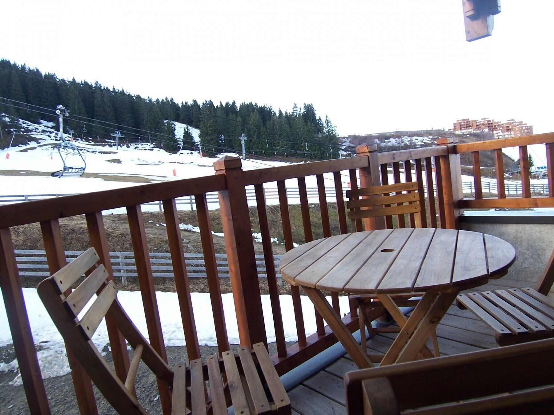 A wooden deck with a round table and chairs, overlooking a snowy landscape with a ski lift in the background.