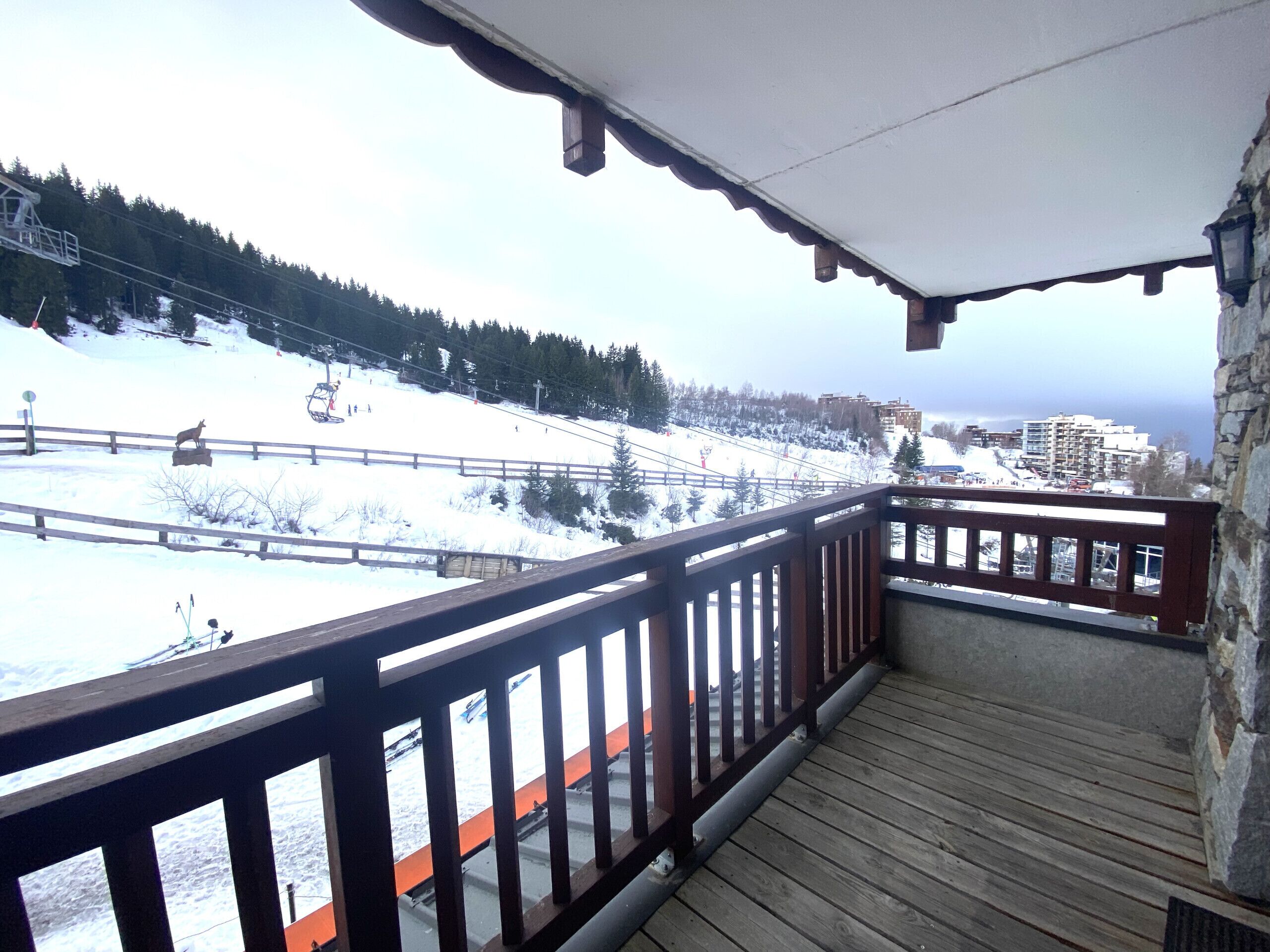 A wooden balcony with a view of a snowy ski slope and buildings in the distance.