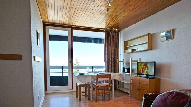 Una sala de estar con techo de madera, un sofá, un televisor, una mesa de comedor con sillas y un ventanal corredizo que da a un balcón.