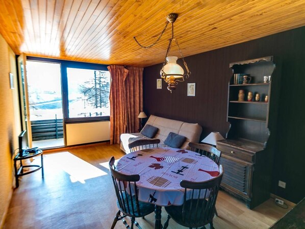 A cozy room with a wooden ceiling, a large window with curtains, a sofa, a dining table with chairs, and a bookshelf.