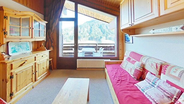 Ein gemütliches Zimmer mit Holzwänden und -decke, einem großen Fenster mit Bergblick, einer Holzkassette mit Schubladen, einem rosa Sofa mit gemusterten Kissen und einem kleinen Tisch.