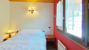 Ein Schlafzimmer mit Bett, Nachttisch mit Lampe, Wandleuchte, Fenster mit Vorhängen und Heizkörper.