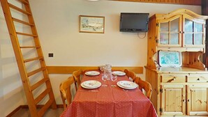 Ein Essbereich mit einem für vier Personen gedeckten Tisch, Holzstühlen, einem Holzkabinett mit Glastüren, einem an der Wand montierten Fernseher und einer an die Wand gelehnten Leiter.