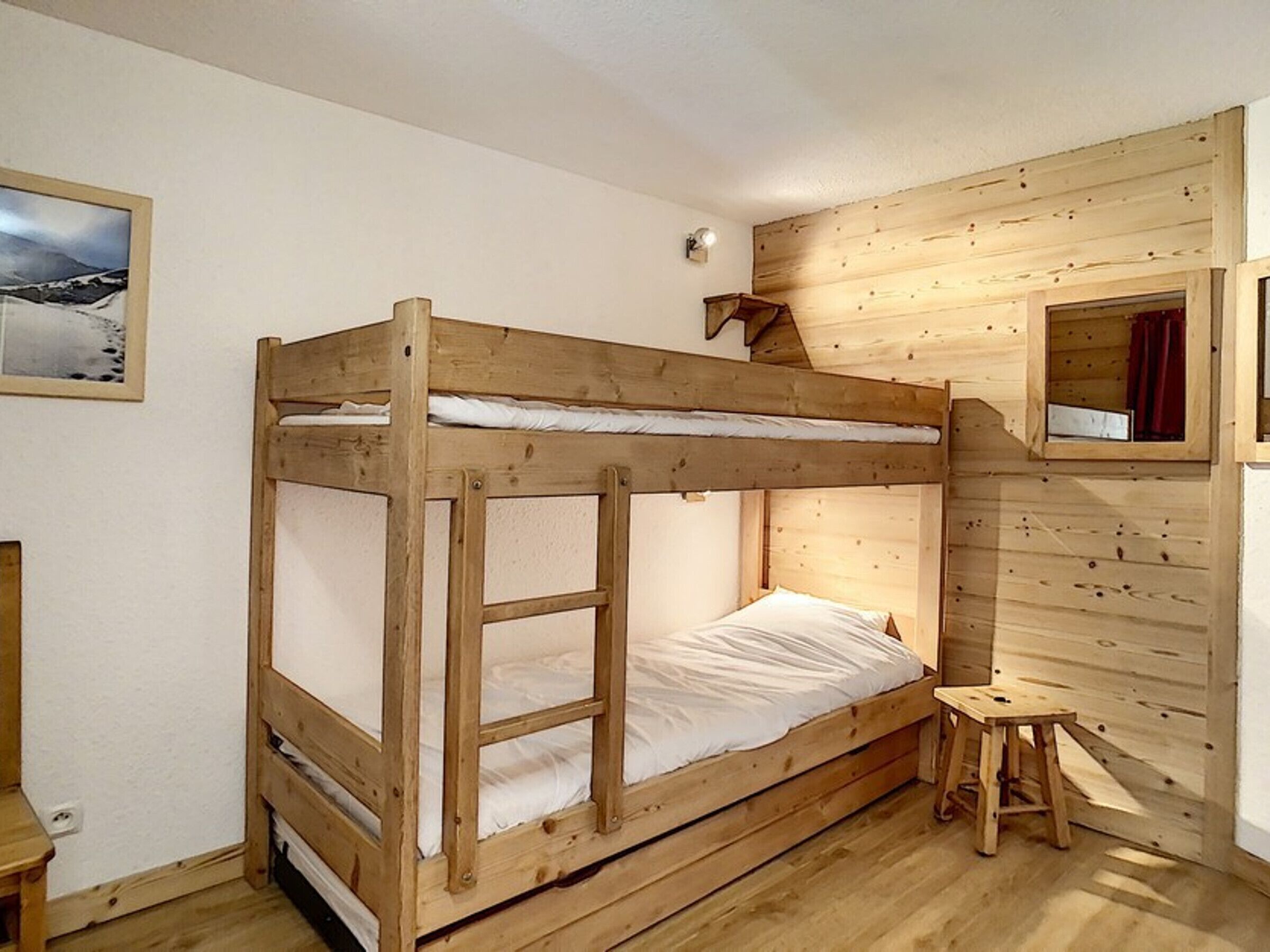 A bunk bed with a wooden frame and white bedding, a small stool, and a mirror on the wall.