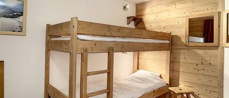 A bunk bed with a wooden frame and white bedding, a small stool, and a mirror on the wall.