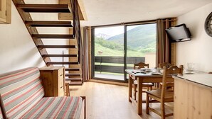 A room with a wooden staircase, a striped sofa, a dining table with chairs, and a large window offering a view of a green landscape. - Modern duplex, ski-in/ski-out, south balcony (Les Menuires)