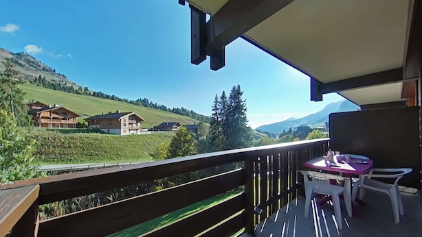 Ein Balkon mit Blick auf ein grünes Tal und Häuser, ein Tisch mit Stühlen und einem hölzernen Geländer.