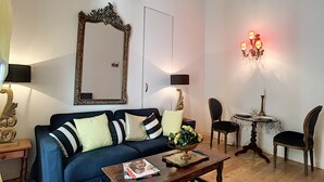 A living room with a blue sofa, a coffee table, a dining table with chairs, a chandelier, and a large mirror on the wall.