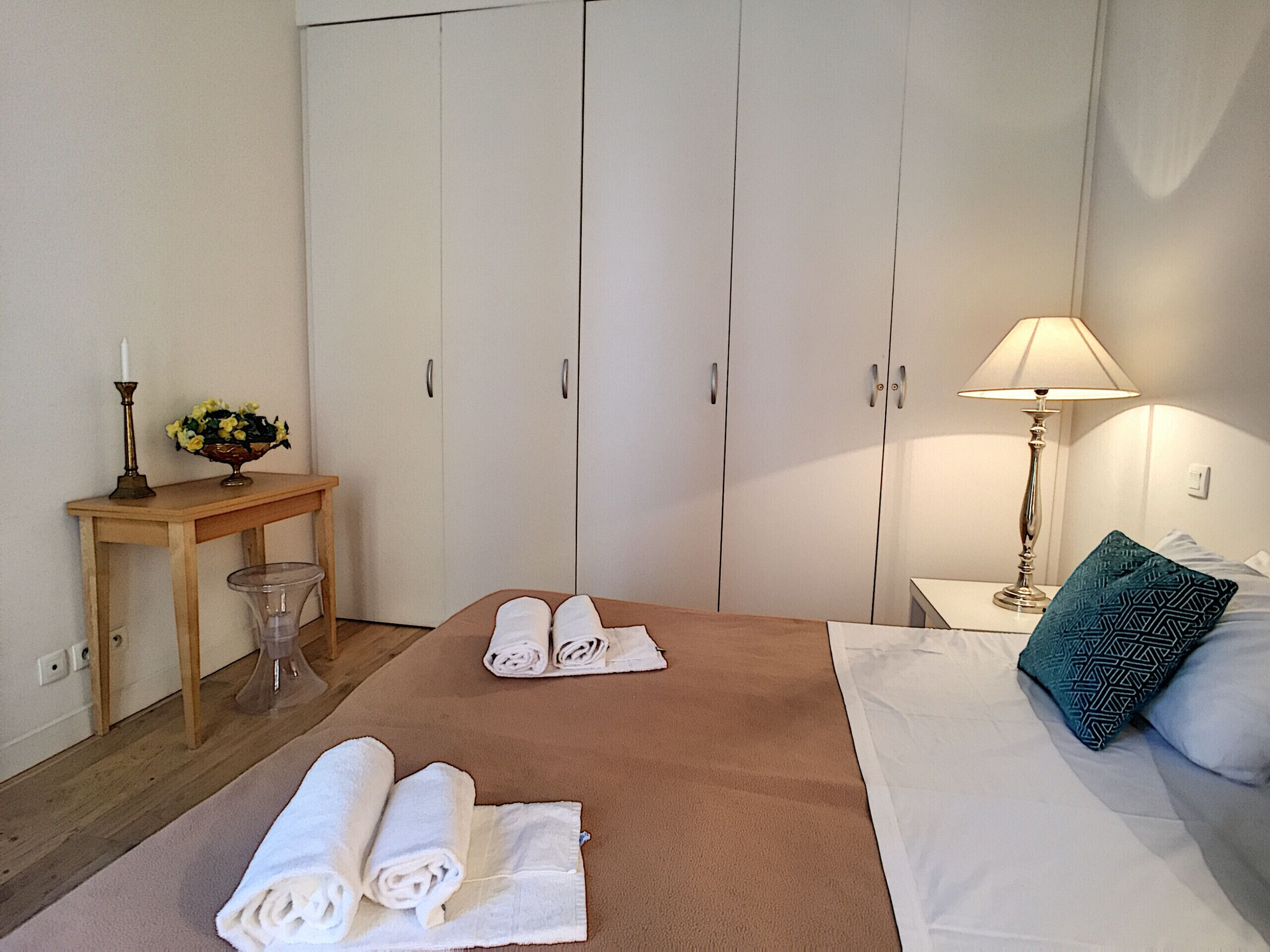 A hotel room with a bed, a nightstand with a lamp, a desk with a vase of flowers, and a wardrobe.