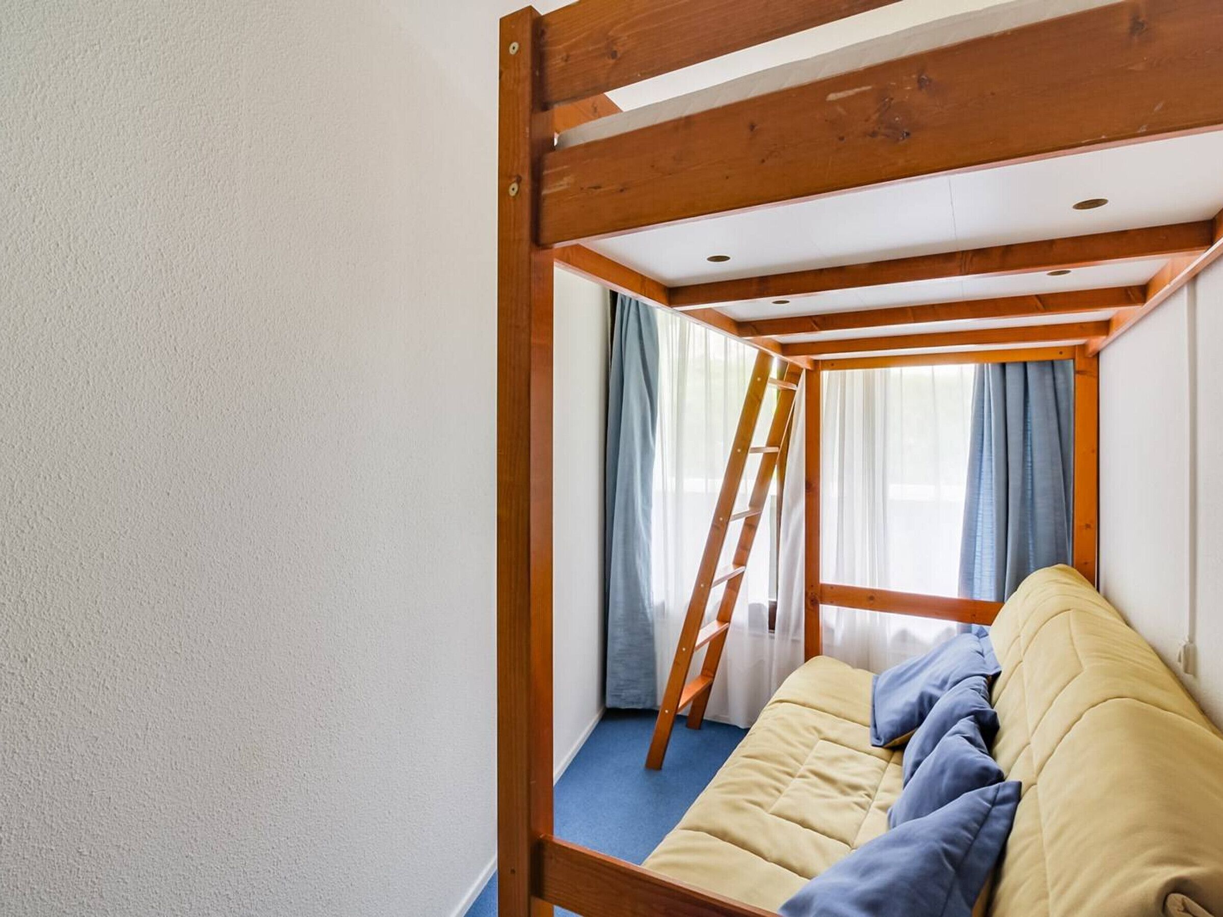 Un lit superposé avec une structure en bois et une échelle, une fenêtre avec des rideaux et un tapis bleu.