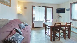 A cozy living room with a sofa, a dining table with chairs, a TV mounted on the wall, and a door leading to an outdoor area. - Cozy 2-Room Apartment with Mountain Views, Near Village Center and Ski Trails - Sleeps 4 (Le Grand-Bornand)