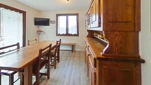 A dining area with a wooden table and chairs, a television mounted on the wall, and a window with curtains. - Cozy 2-Room Apartment with Mountain Views, Near Village Center and Ski Trails - Sleeps 4 (Le Grand-Bornand)