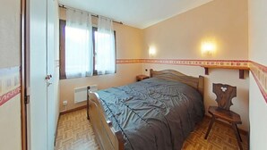 A bedroom with a wooden bed, a nightstand, a window with curtains, a radiator, and a wall-mounted light fixture. - Cozy 2-Room Apartment with Mountain Views, Near Village Center and Ski Trails - Sleeps 4 (Le Grand-Bornand)