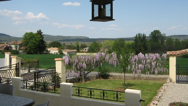 Un patio con mobiliario exterior, vista a un jardín con flores moradas y un paisaje distante con casas y colinas.
