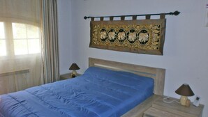 Un dormitorio con una cama, dos mesitas de noche, un adorno en la pared y una ventana con cortinas.
