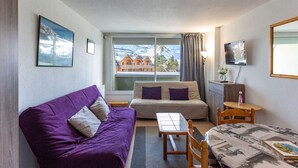 A cozy room with a purple sofa, a beige sofa, a small table, and a bed with a patterned bedspread. - Cozy 5-person studio near slopes, parking included (La Mongie)