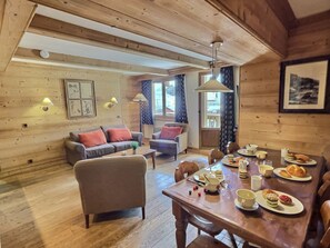 A cozy wooden interior with a dining table set for breakfast, a sofa, and a window with curtains.