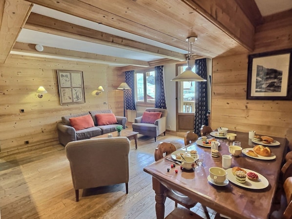 A cozy wooden interior with a dining table set for breakfast, a sofa, and a window with curtains.