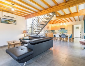 Un intérieur moderne avec un canapé en cuir noir, des poutres en bois, un escalier et un coin cuisine.