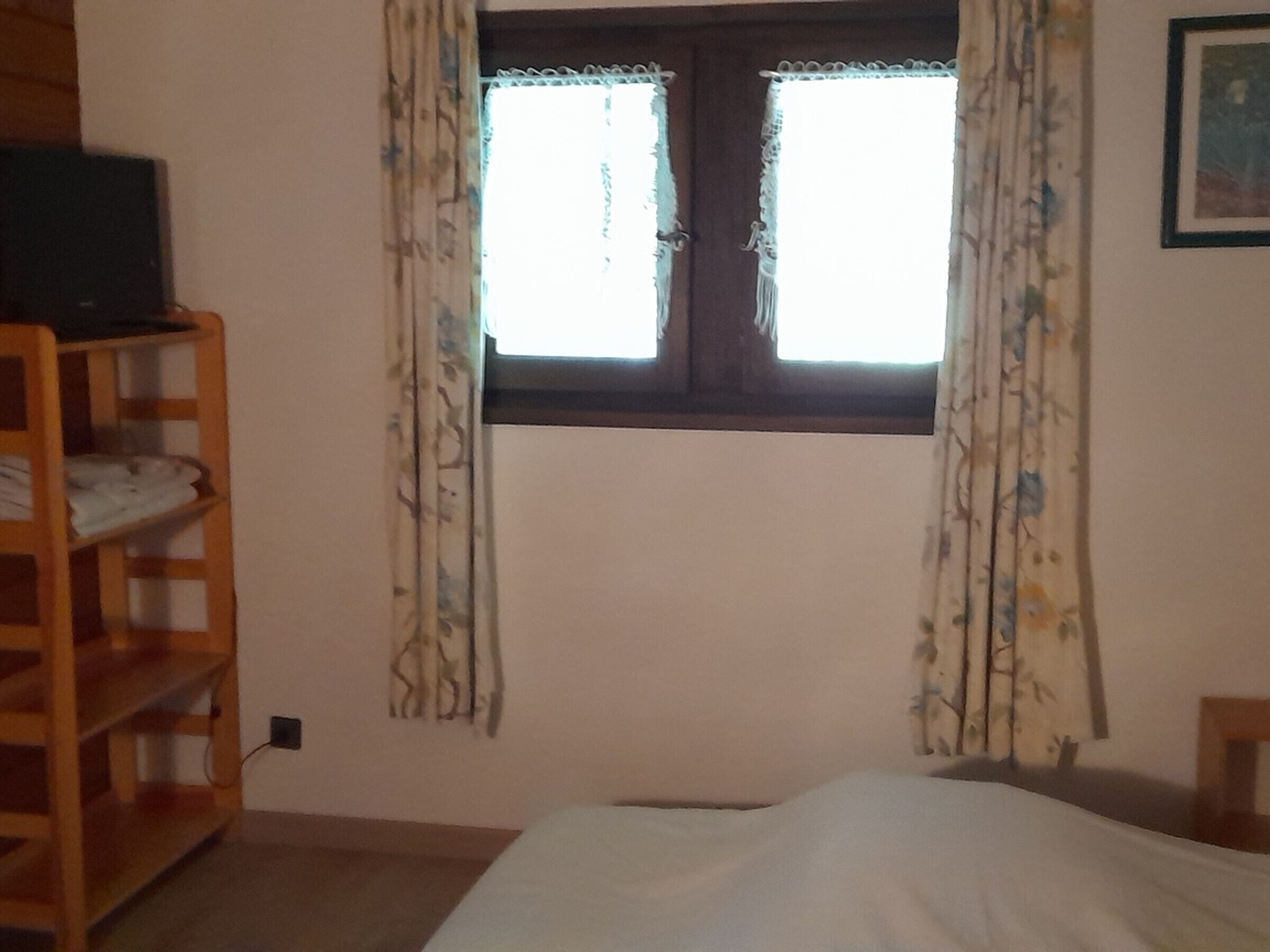 A bedroom with a bed, a wooden shelf unit, a window with curtains, and a wall-mounted picture.