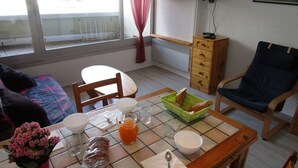 Un comedor con una mesa dispuesta para el desayuno, incluyendo pan, un tazón y un vaso de jugo de naranja. Se observa una silla, una mesita auxiliar y una ventana con cortinas.