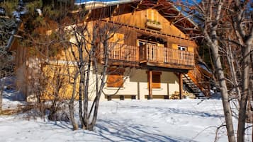 A wooden chalet with a balcony, surrounded by snow and bare trees.