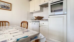 A compact kitchen with white cabinetry, a built-in oven, and a dining table with a patterned tablecloth. - 2-bedroom apartment near sea and center, includes parking (Cucq-Stella Plage)