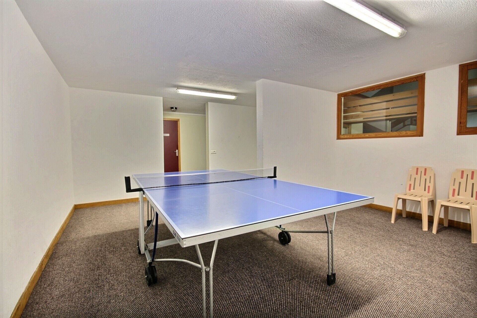 Une pièce avec une table de ping-pong, deux chaises, un sol recouvert de moquette, des murs blancs et une porte au fond.