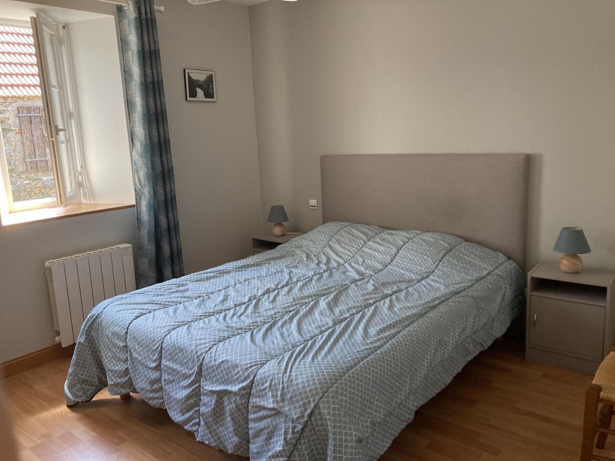 Un dormitorio con cama, dos mesitas de noche, ventana con cortinas, radiador y un cuadro en la pared.
