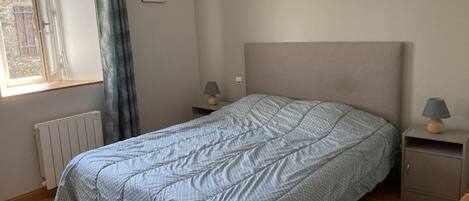Un dormitorio con cama, dos mesitas de noche, ventana con cortinas, radiador y un cuadro en la pared.
