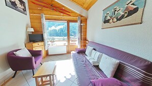 Un salon chaleureux avec un canapé violet, une table basse en bois et un téléviseur posé sur un meuble en bois. Une grande fenêtre offre une vue sur un paysage enneigé.