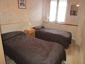 Une chambre avec deux lits simples, une table de chevet avec une lampe, un radiateur et une fenêtre avec des rideaux.