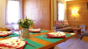 Une pièce chaleureuse avec une table à manger en bois, dressée pour deux personnes, un canapé agrémenté d’un coussin coloré et une lampe fixée au mur.