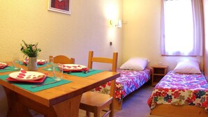 Une petite chambre accueillante, avec une table en bois dressée pour deux personnes, deux lits simples avec une literie à motifs floraux, une petite table de chevet et une fenêtre avec des rideaux.