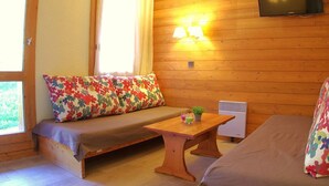 Une pièce chaleureuse avec un mur en bois, un canapé agrémenté de coussins à motifs floraux colorés, une petite table avec une plante et un téléviseur à écran plat fixé au-dessus d’un panneau en bois.