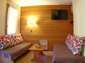 A cozy room with wooden panel walls, a flat-screen TV mounted above a wooden table, and two sofas with floral-patterned cushions.