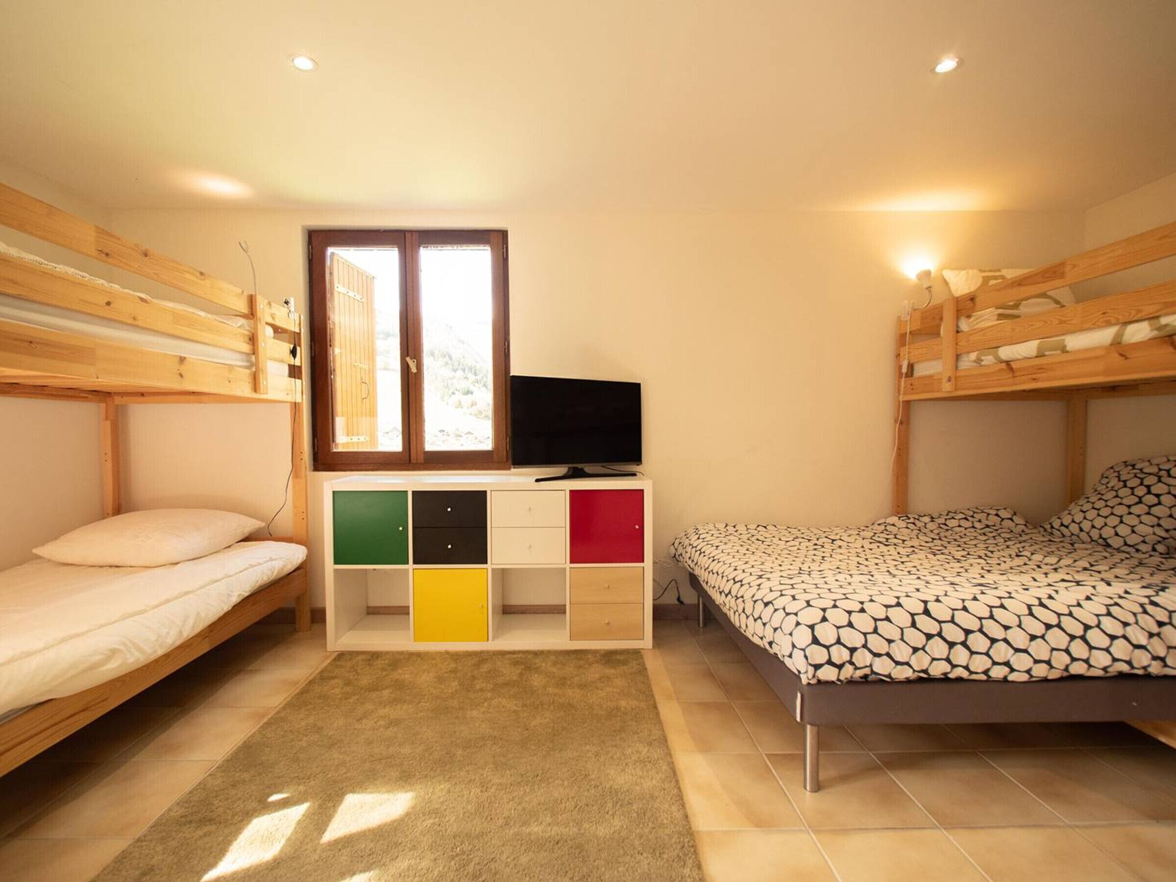 A room with two bunk beds, a TV on a wooden stand, a window, and a storage unit with colorful drawers.