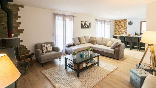 A modern living room with a sofa, armchair, and coffee table. There is a fireplace, a dining area, and a kitchen in the background. - Renovated 7-room chalet in Valmorel, 15 people, parking, terrace (Valmorel)