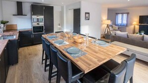 A modern kitchen with dark cabinets, a wooden dining table set with plates and glasses, and a living area with a sofa and lamp. - Renovated 7-room chalet in Valmorel, 15 people, parking, terrace (Valmorel)