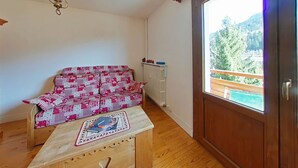 A room with a wooden floor, a patterned sofa, a small table with a book, a radiator, and a window with a view of greenery.