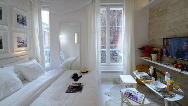 Ein Schlafzimmer mit einem Bett, einem Nachttisch mit Büchern und einer Kamera, einem Fenster mit Blick auf Bäume und einem an der Wand montierten Fernseher.
