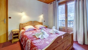 Una cama de madera con una colcha roja estampada, dos mesitas de noche y una ventana con cortinas.