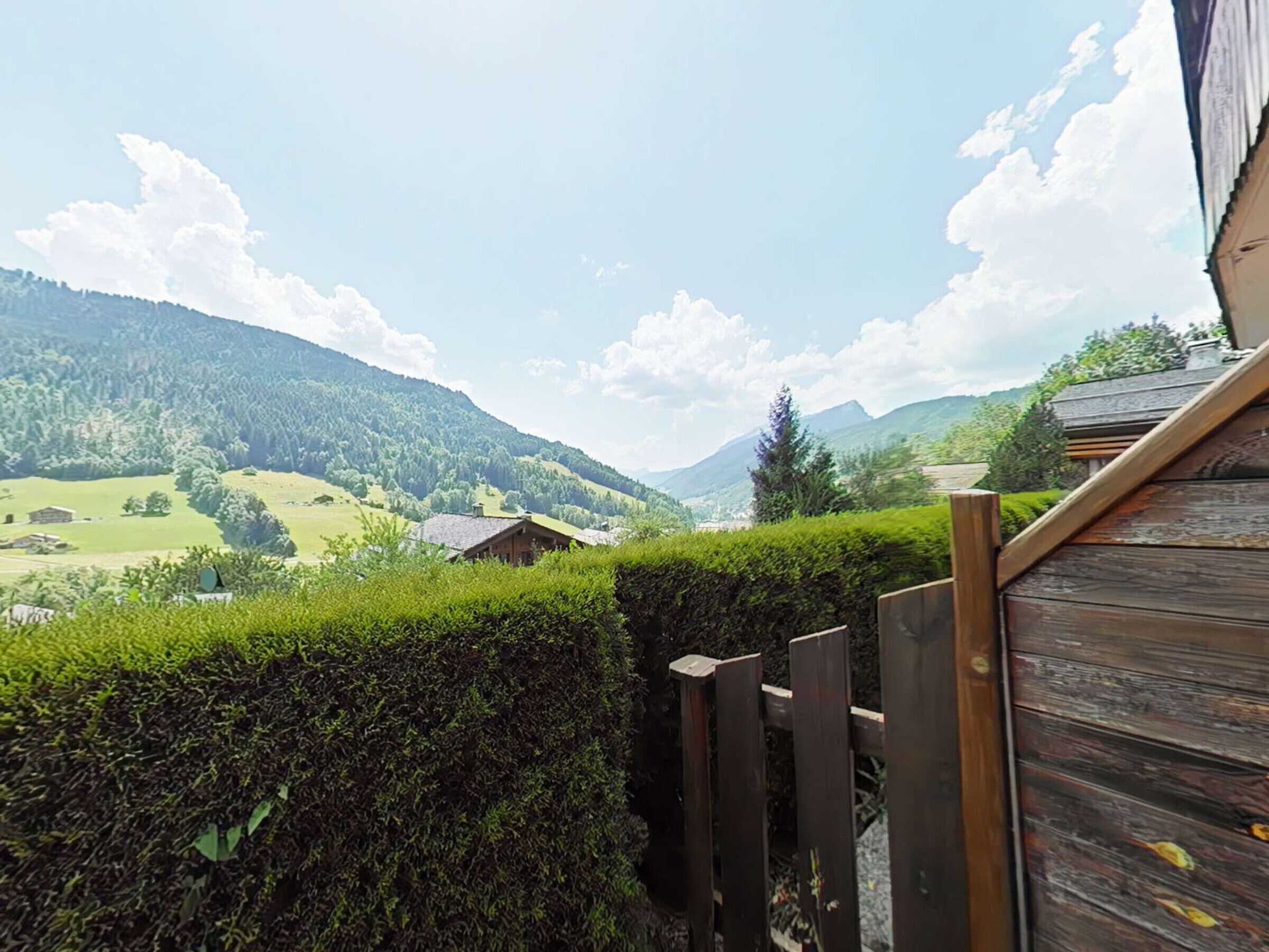 A wooden deck with a view of a mountainous landscape, including a house and a hedge.