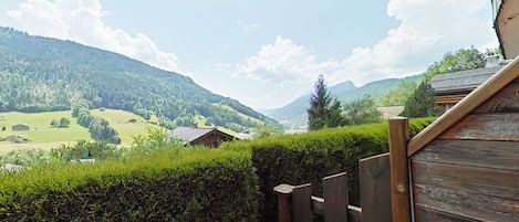 A wooden deck with a view of a mountainous landscape, including a house and a hedge.