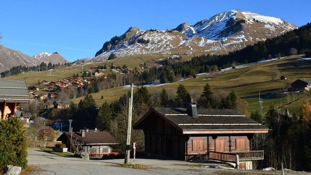 Un village de montagne aux maisons traditionnelles en bois, avec un ciel dégagé et des sommets enneigés en toile de fond.