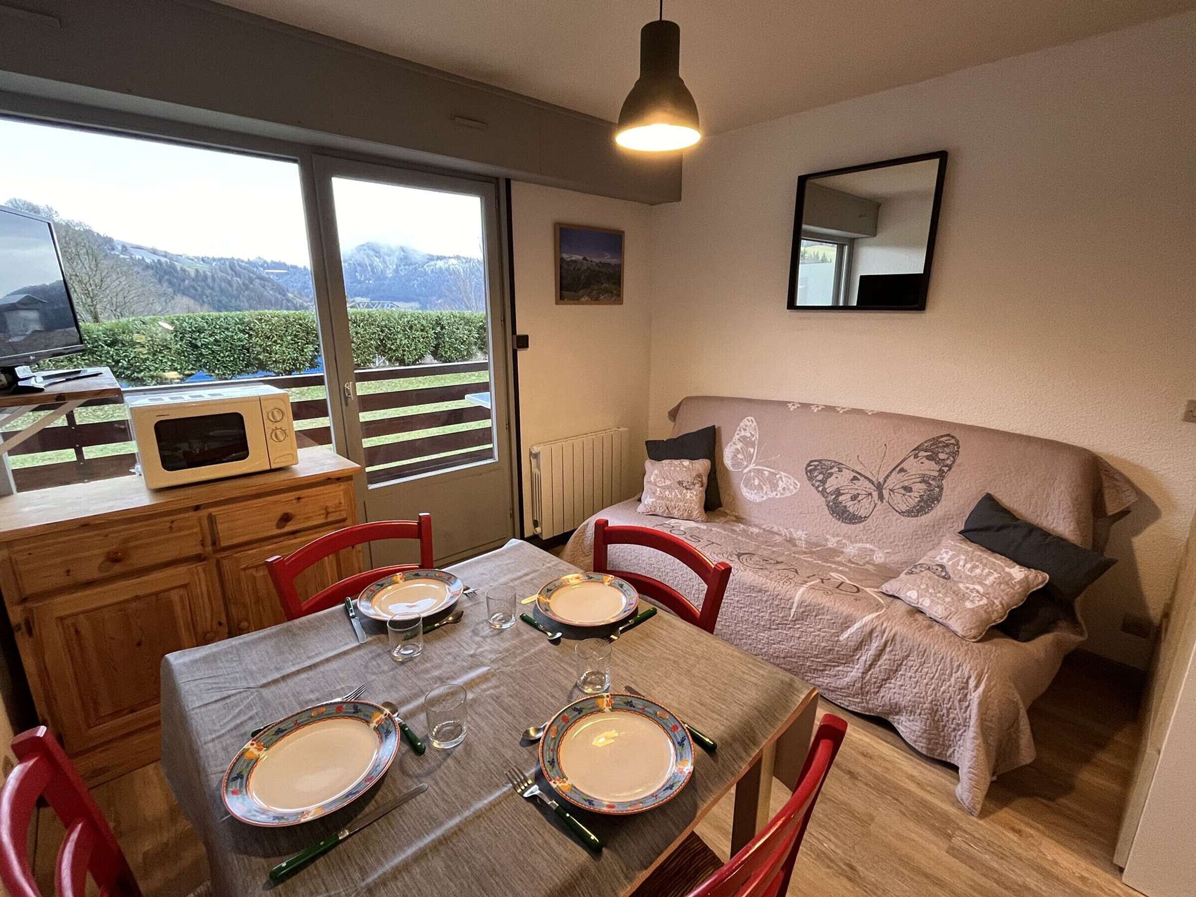 A dining area with a table set for two, a sofa with butterfly designs, and a view of mountains through a large window.